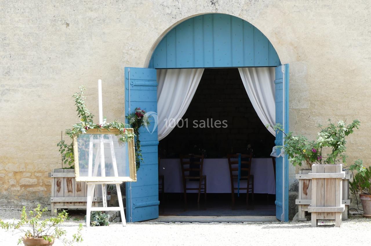 Porte bleue ouverte sur une salle avec des chaises, encadrée de plantes en pots et de rideaux blancs.