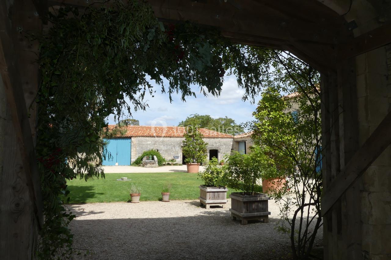 Vue d'une cour intérieure avec bâtiments en pierre, toits en tuiles, pelouse et jardinières sous une arche en bois.