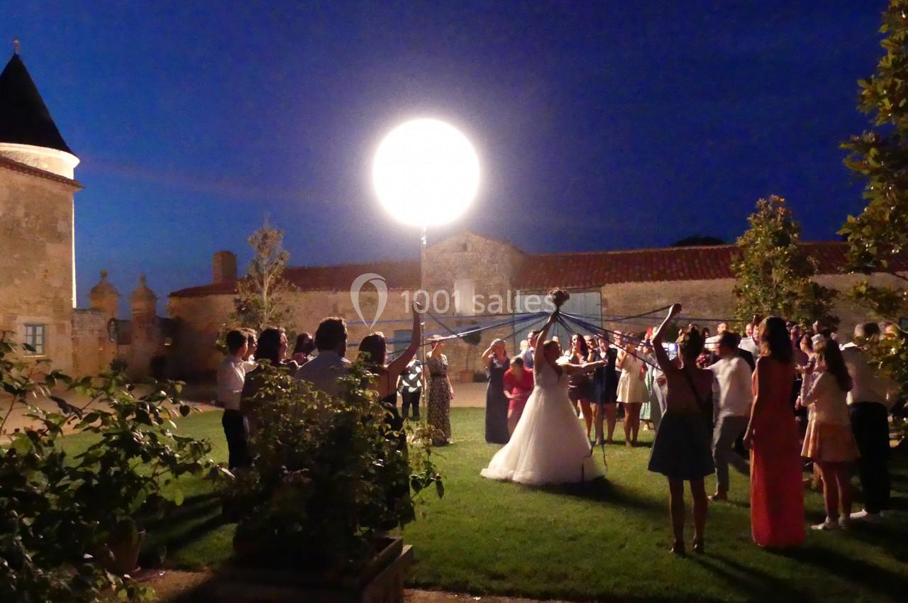 Groupe de personnes entourant un couple de mariés dans un jardin éclairé par un grand projecteur, en soirée.