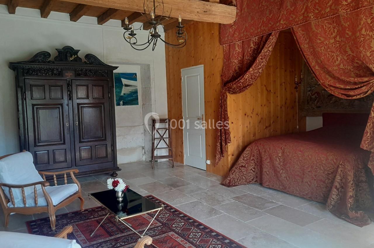 Chambre avec lit à baldaquin rouge, armoire en bois sculpté, fauteuils et table basse sur tapis, poutres apparentes.