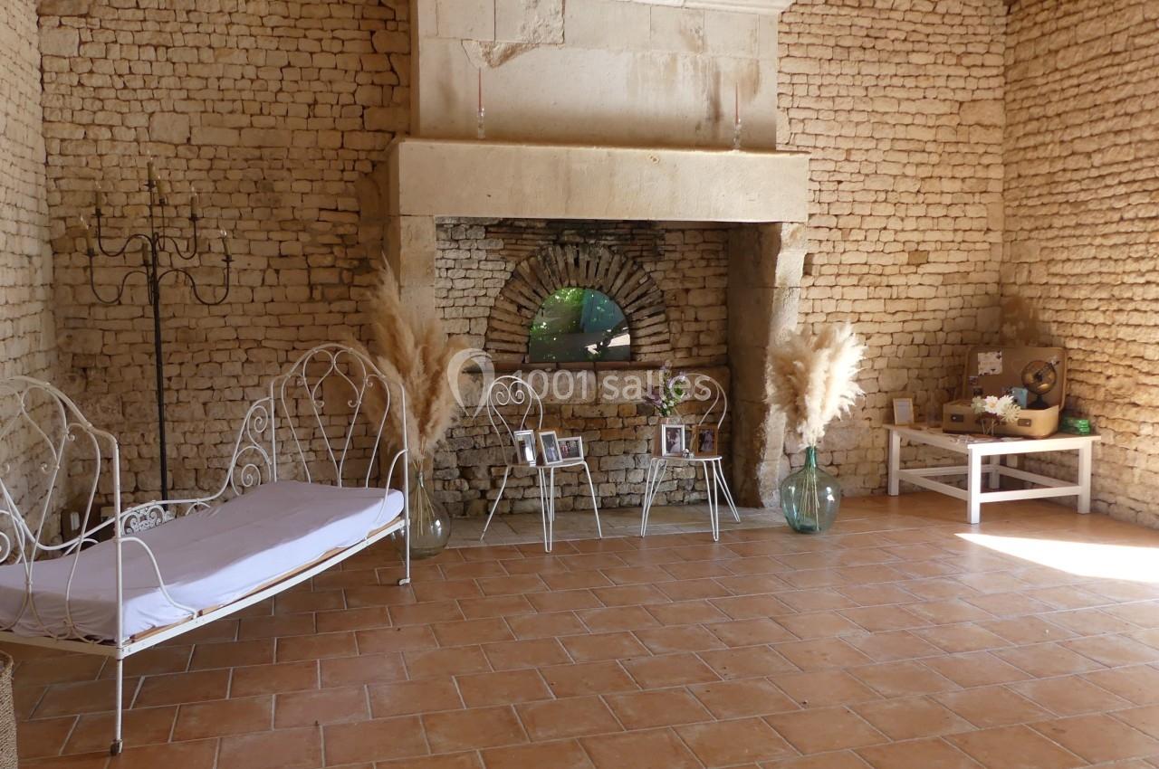 Salle en pierre avec cheminée ancienne, canapé en fer forgé, décorations végétales et lumière naturelle.