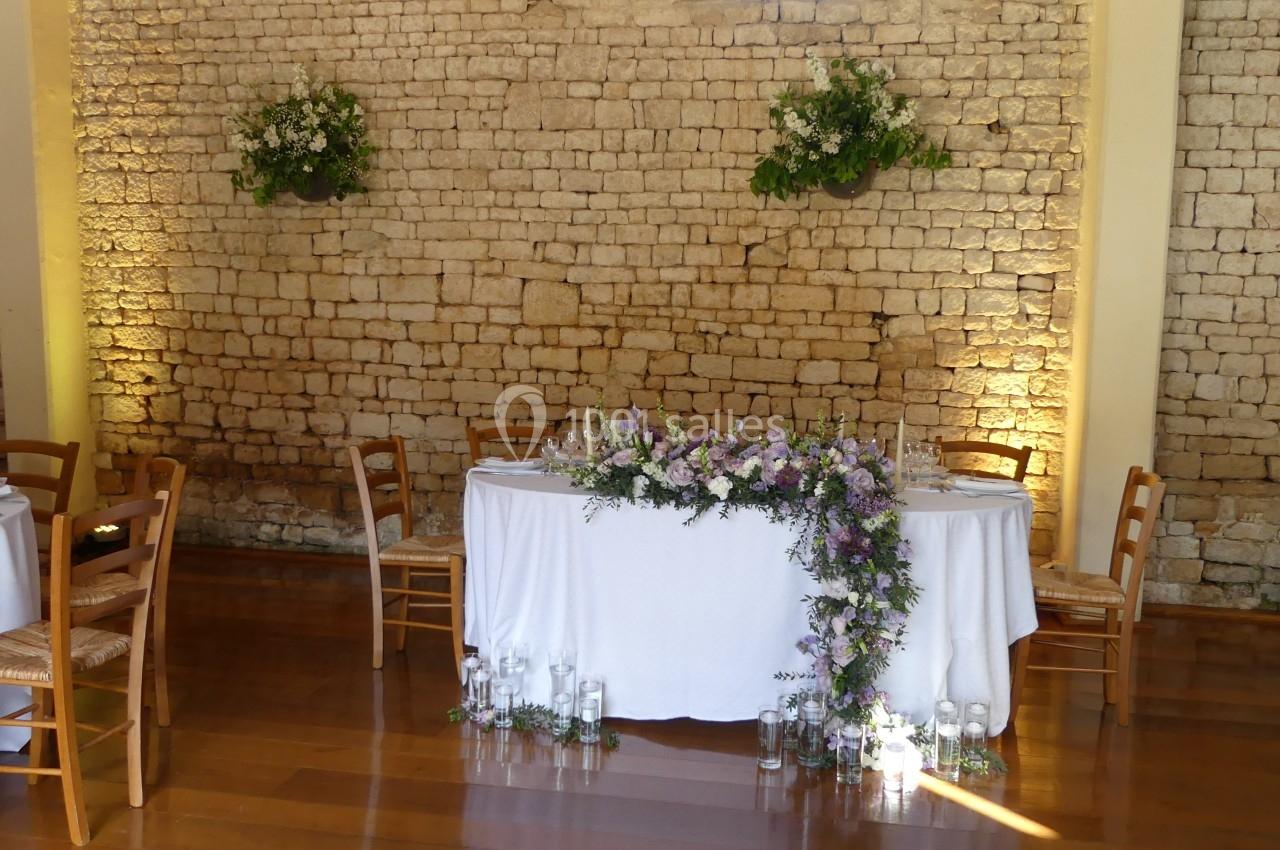 Table décorée pour un événement, ornée de fleurs violettes et blanches, devant un mur en pierre éclairé.