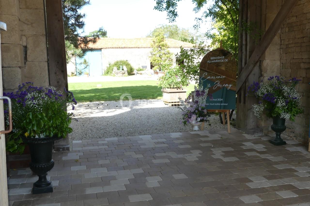 Entrée pavée d'une cour avec des fleurs en pots, une enseigne en bois et un jardin en arrière-plan.