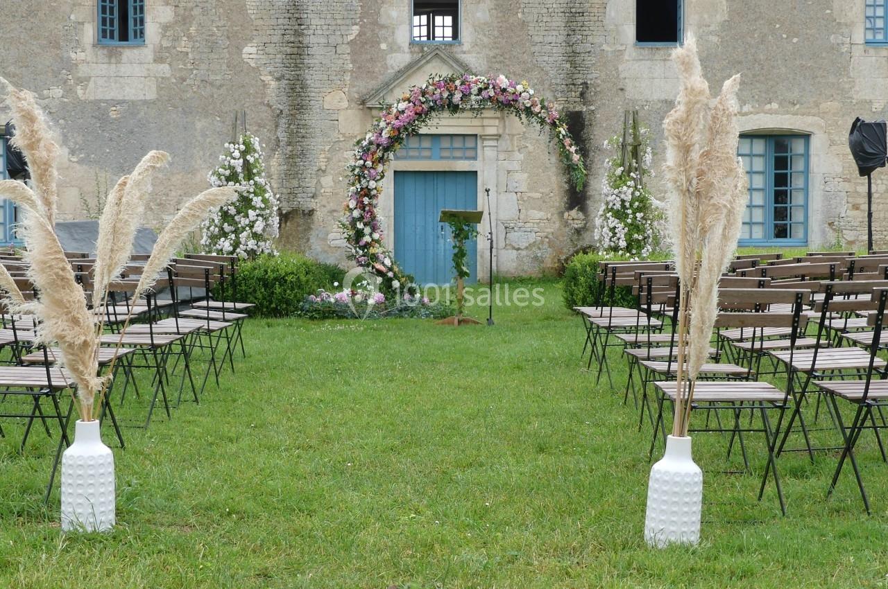 Allée centrale d'une cérémonie en plein air, décorée d'herbes de la pampa et d'une arche fleurie devant un bâtiment ancien.