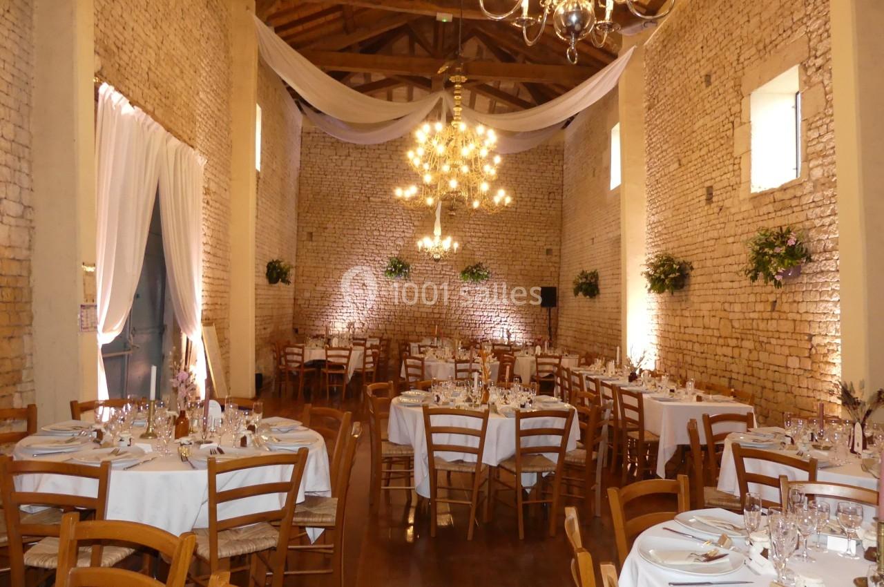 Salle de réception en pierre avec tables rondes dressées, chaises en bois, lustres suspendus et décorations florales.