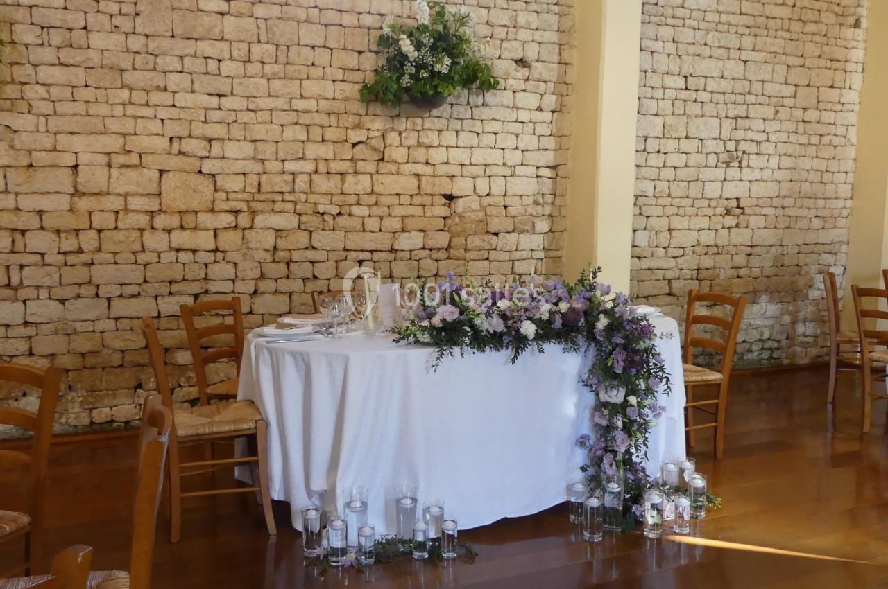 Table décorée pour un mariage avec nappes blanches, fleurs violettes et blanches, bougies et mur en pierre en arrière-plan.