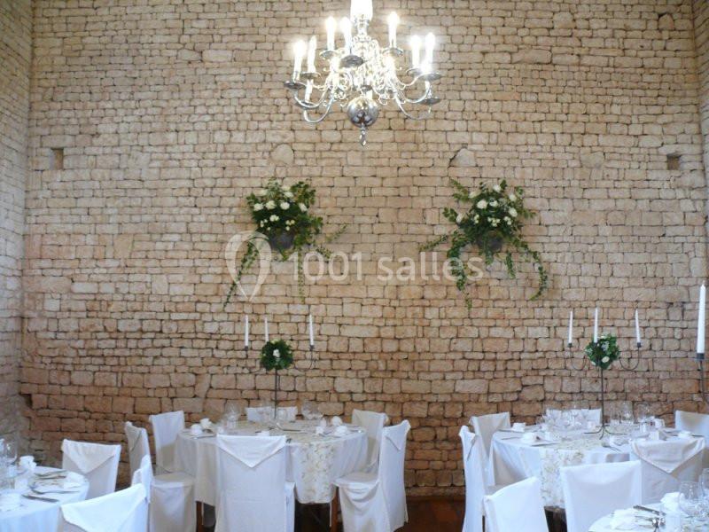 Salle de réception avec murs en pierre, tables dressées avec nappes blanches et chandelier suspendu.