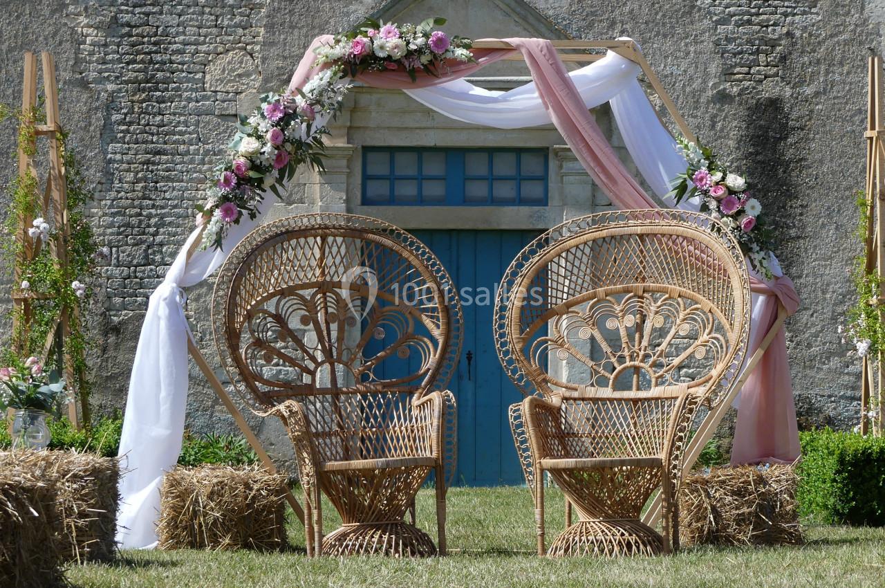 Deux fauteuils en rotin devant une arche décorée de fleurs et de tissus, installée près d'une porte bleue sur un mur en…