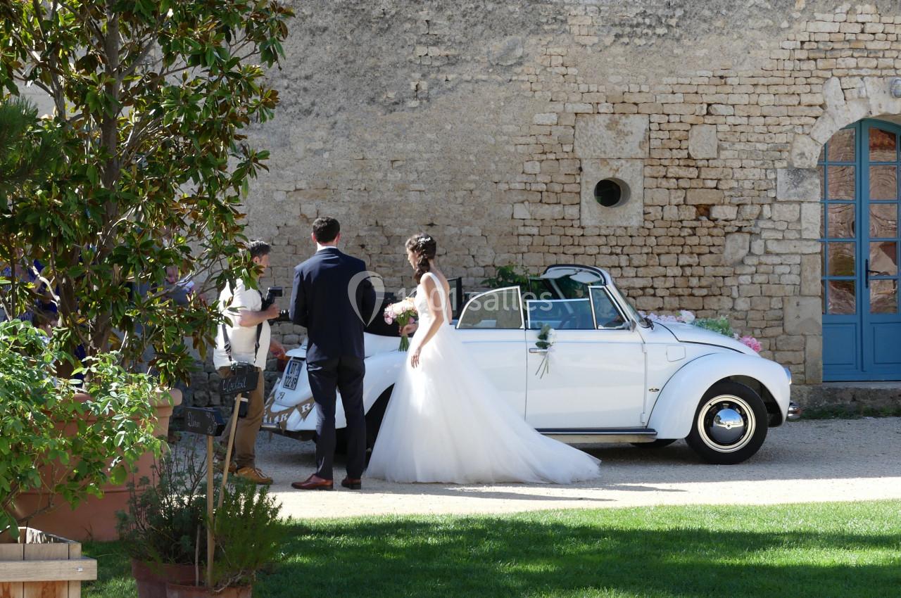 Un couple de mariés se tient près d'une voiture blanche décapotable décorée, devant un mur en pierre.