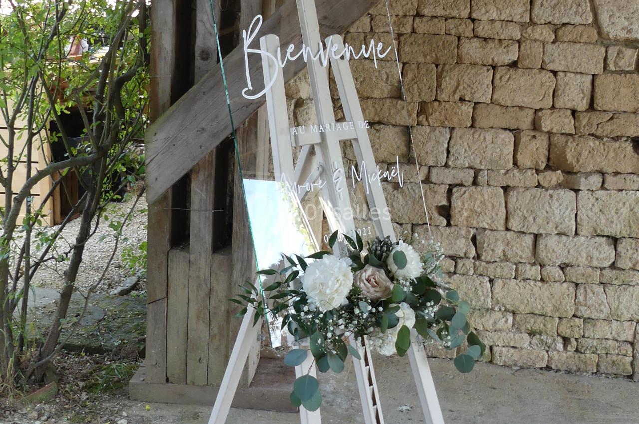 Panneau de bienvenue pour un mariage, en verre transparent, décoré de fleurs blanches et vertes, posé sur un chevalet.