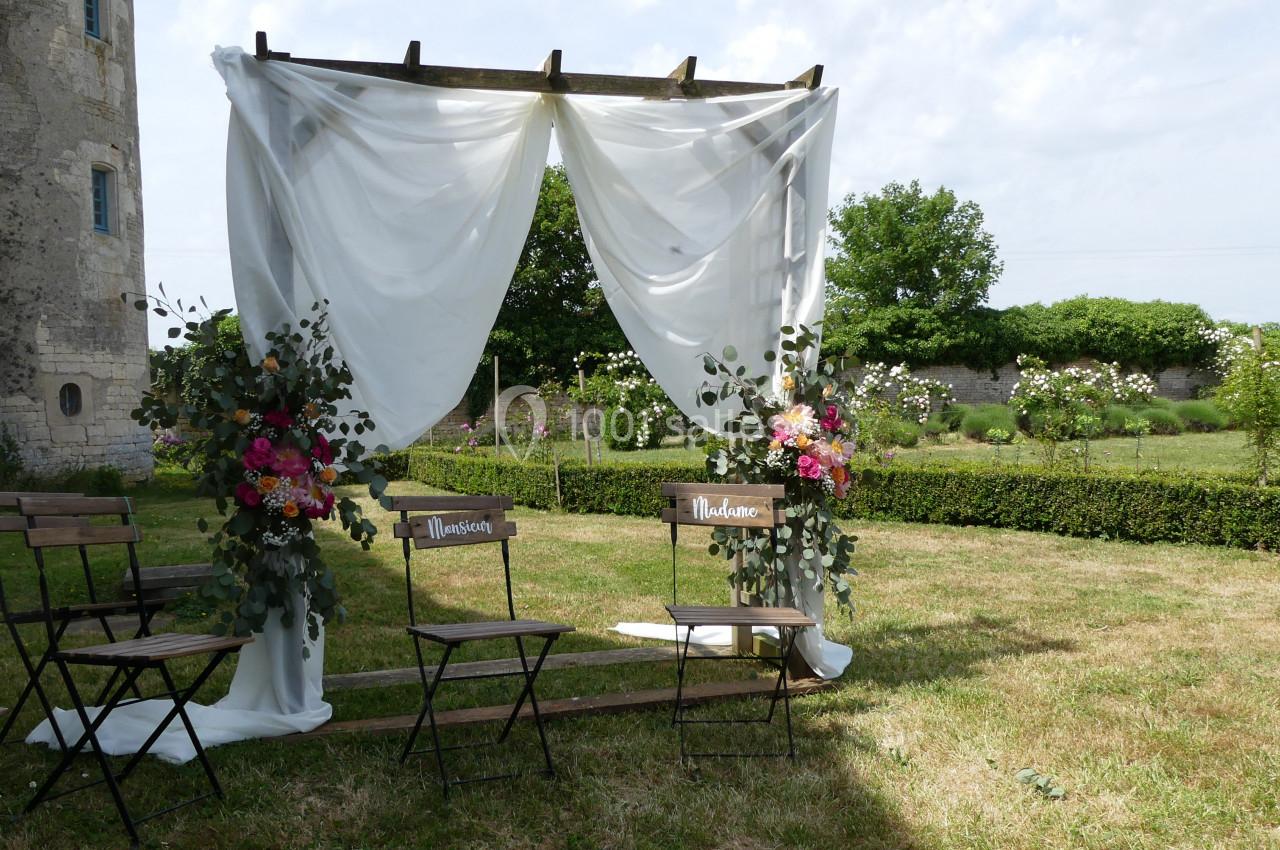 Arche de mariage en bois décorée de voilages blancs et de fleurs colorées, avec deux chaises sur une pelouse.