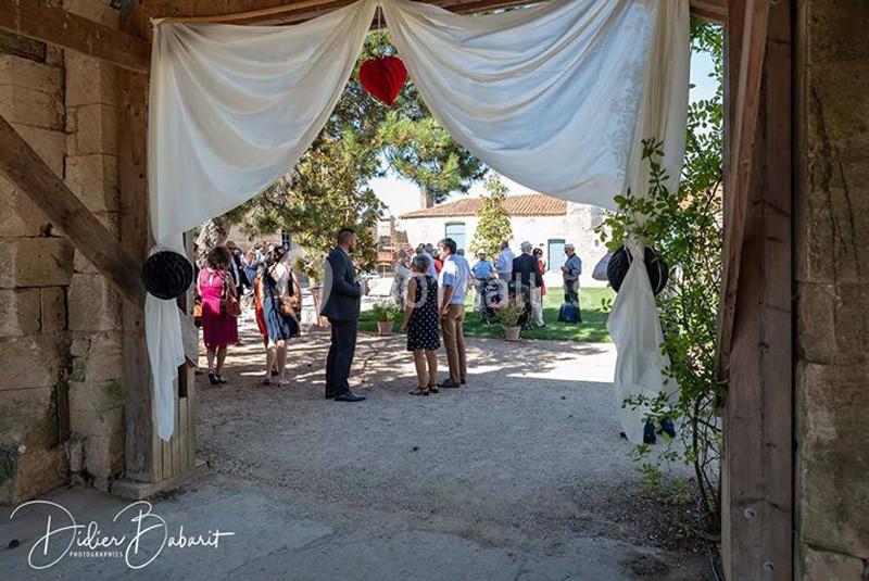 Des invités discutent dans une cour extérieure ensoleillée, vue à travers une arche décorée de rideaux blancs.