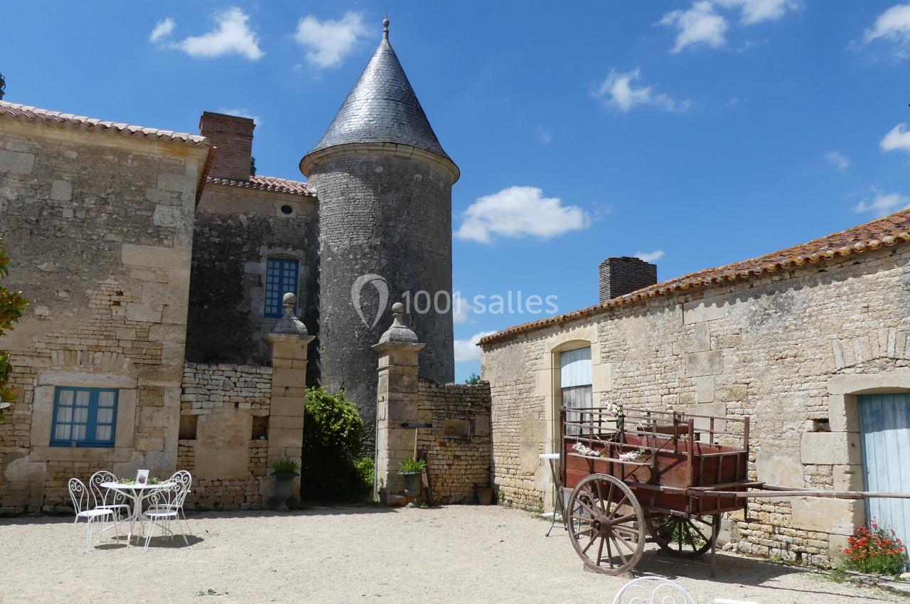 Cour intérieure d'un bâtiment en pierre avec une tour, une charrette en bois et des tables blanches sous un ciel bleu.