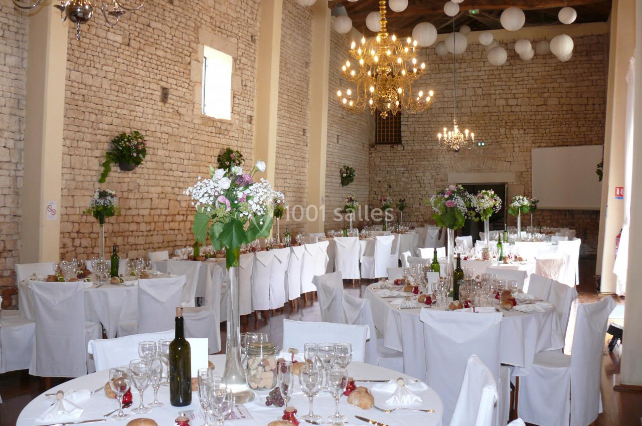 Salle de réception décorée avec des tables rondes dressées, des chaises blanches et des fleurs suspendues.
