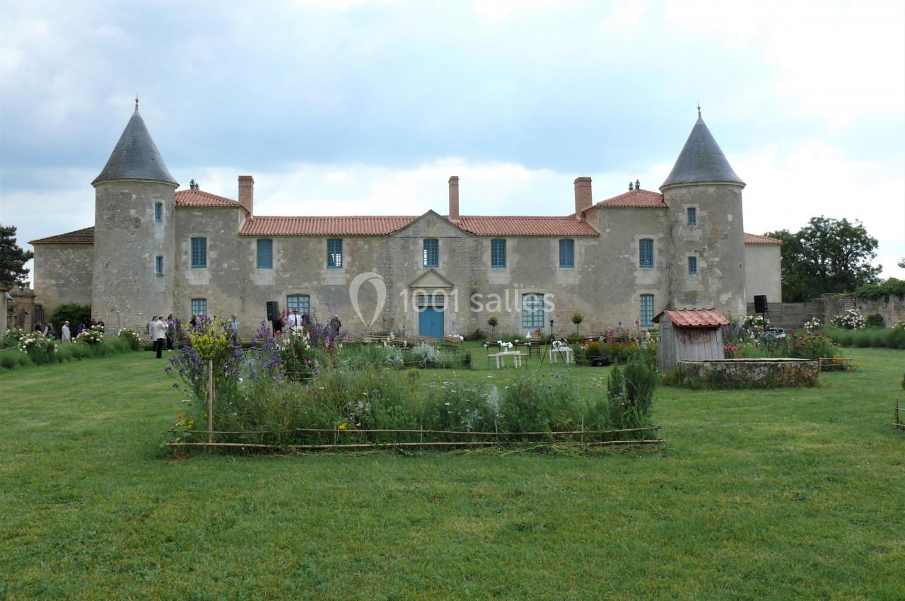 Façade d'un château en pierre avec deux tours, entouré d'un jardin fleuri et d'une pelouse verte.
