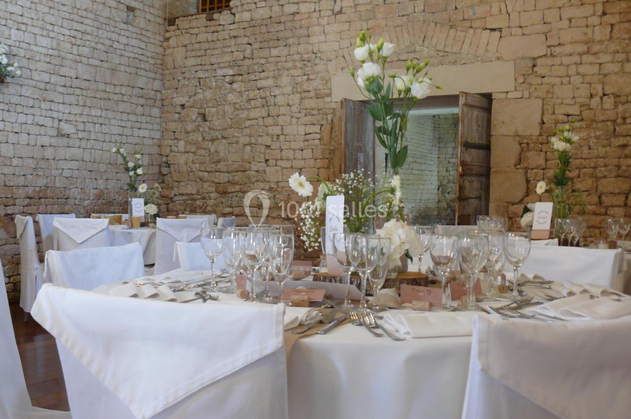 Table de réception élégamment dressée avec nappes blanches, verres, couverts et décoration florale dans une salle en pierre.
