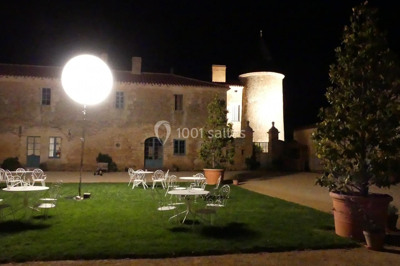 Cour éclairée la nuit avec des tables et chaises en fer forgé sur une pelouse, devant un bâtiment en pierre.