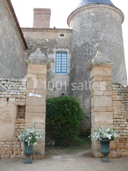 Entrée en pierre d'un bâtiment ancien avec deux piliers décorés de fleurs blanches et une tour en arrière-plan.