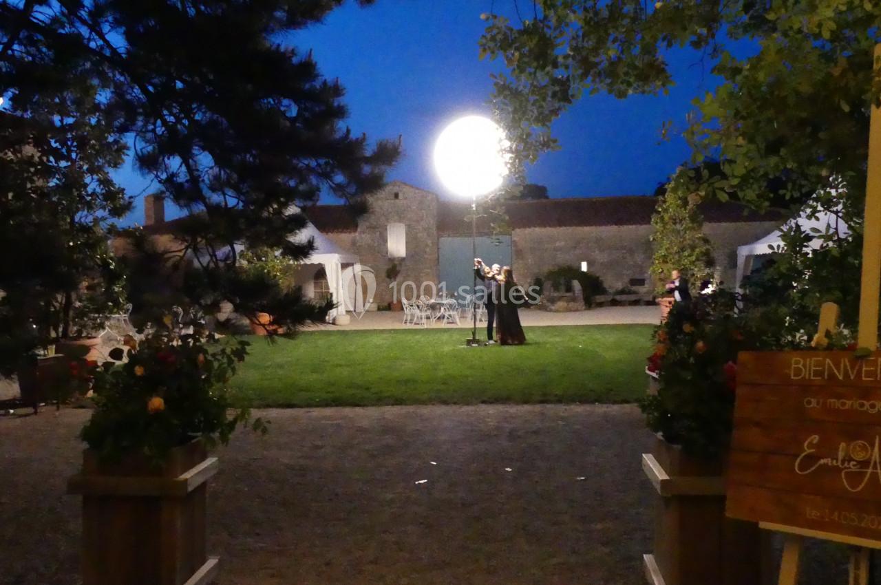 Un couple danse sous une lumière ronde dans un jardin éclairé, entouré de bâtiments en pierre au crépuscule.