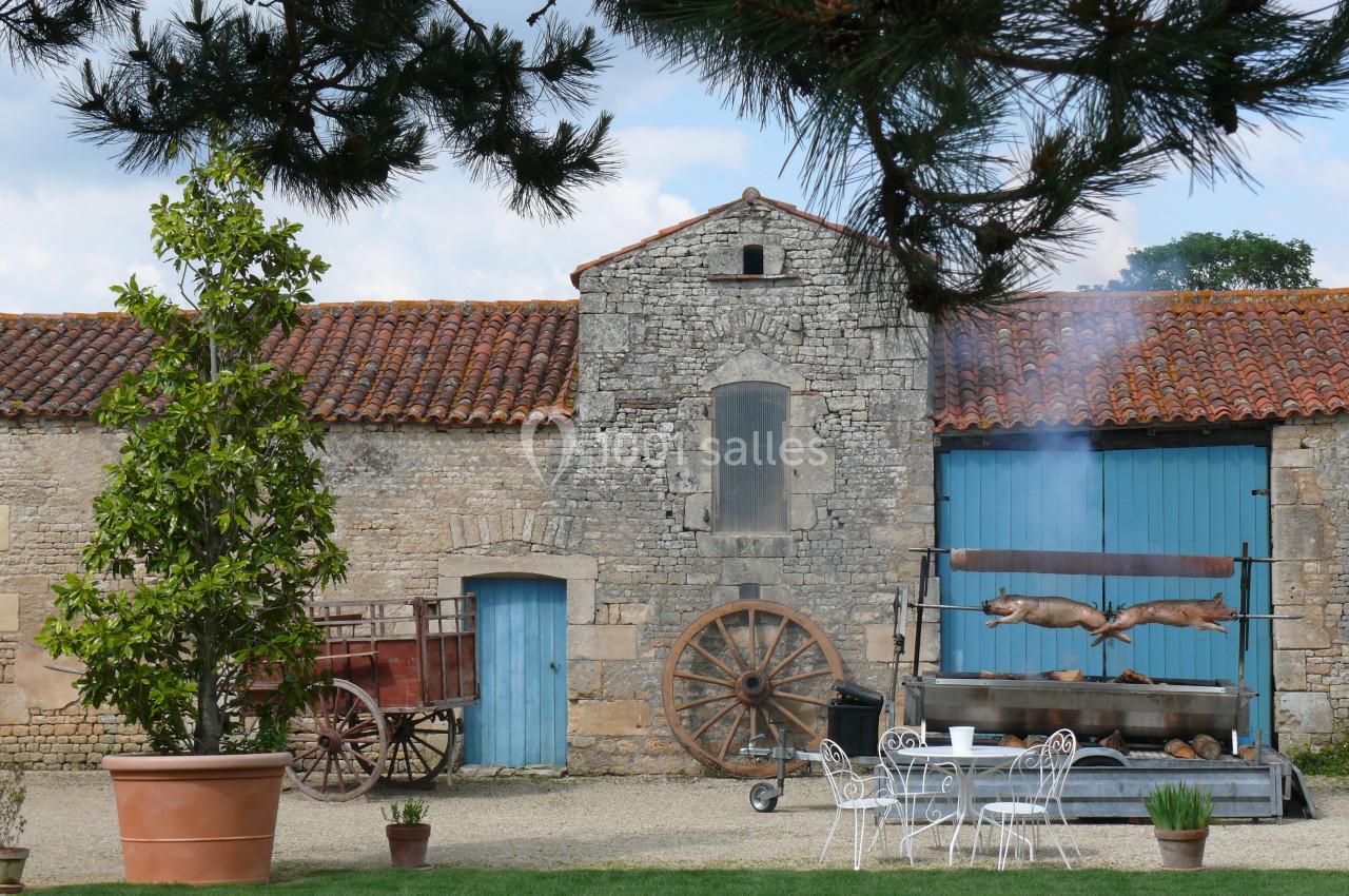 Cour d'une ferme avec murs en pierre, charrette ancienne, table en fer forgé et rôtissoire en fonctionnement.