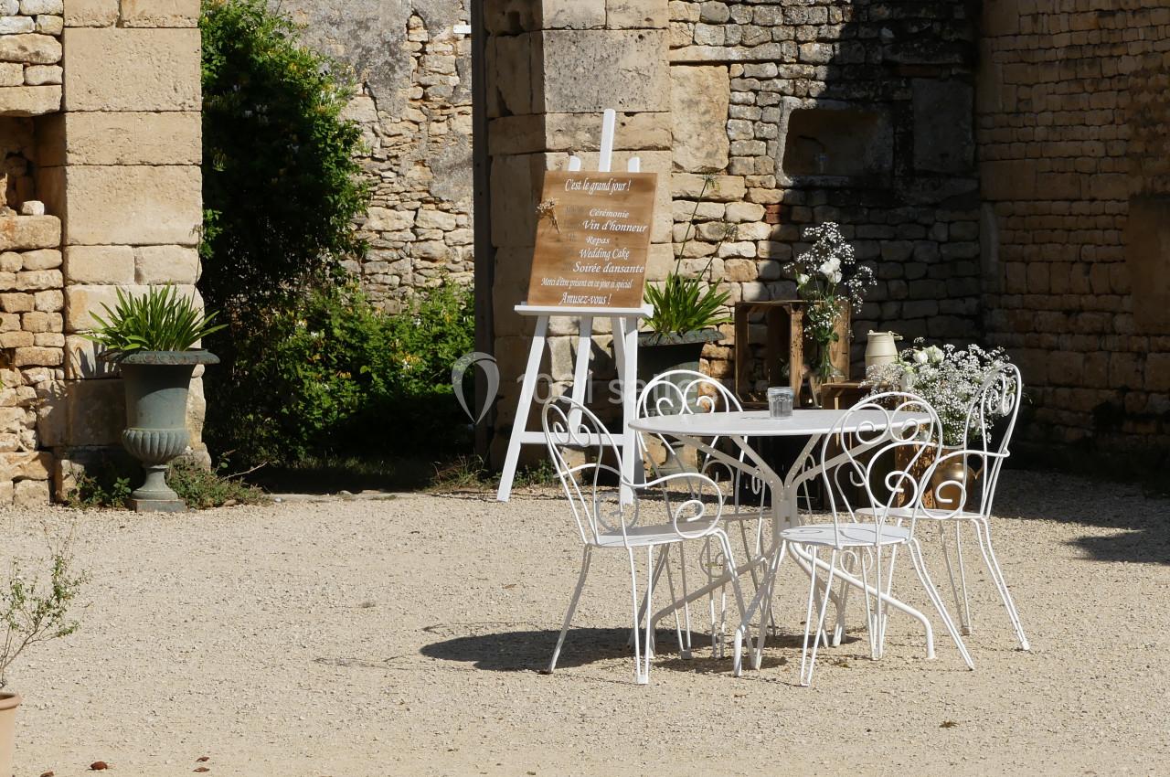 Table et chaises blanches en fer forgé disposées sur une terrasse en gravier, entourées de plantes et de murs en pierre.