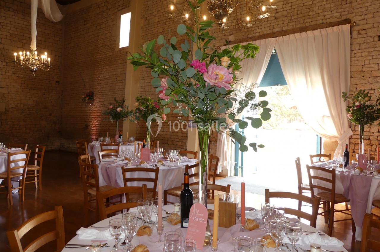 Salle de réception décorée avec des tables dressées, nappes roses, bouquets de fleurs et lumière naturelle entrant par une…