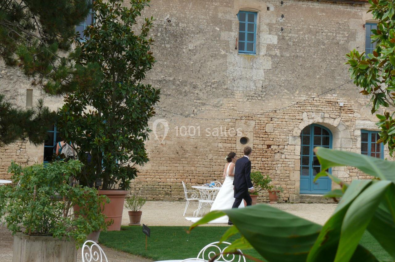 Un couple marche dans la cour d'une maison en pierre avec des fenêtres bleues, entourée de végétation.