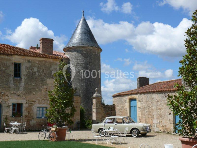 Cour d'une bâtisse en pierre avec une tour, des tables en fer forgé, et une voiture ancienne stationnée.