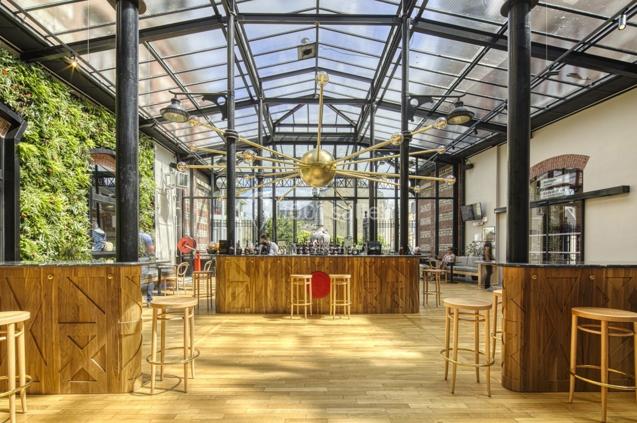 Intérieur lumineux d'un bar avec un toit en verre, un comptoir central en bois et des tabourets autour.