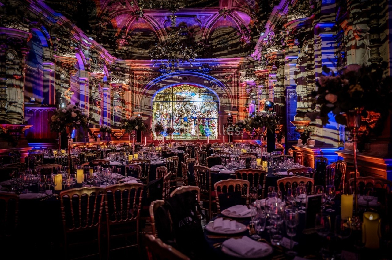 Salle de réception décorée avec des tables dressées, éclairages colorés et vitrail en arrière-plan.