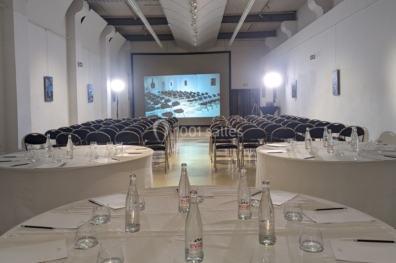 Salle de conférence avec des rangées de chaises alignées devant un écran de projection, tables rondes au premier plan.