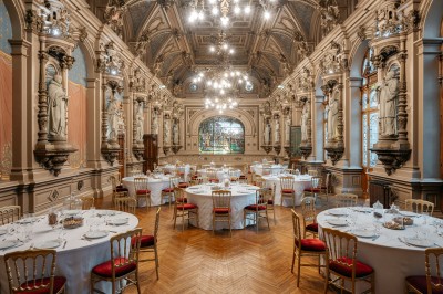 Salle de réception élégante avec tables rondes dressées, décorée de statues, fresques et lustres imposants.