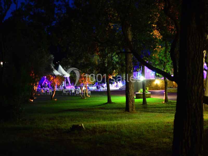 Espace extérieur éclairé la nuit avec des arbres et une tente blanche au loin, entouré de lumières colorées.