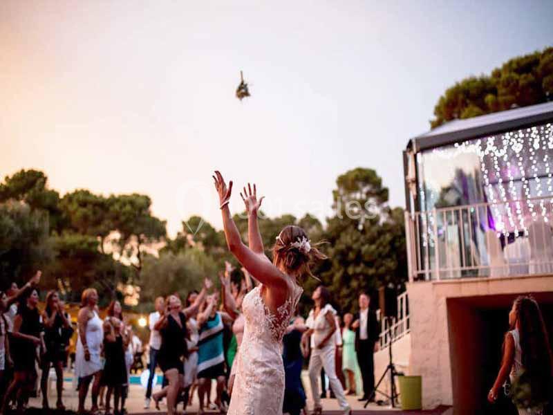 Une mariée lance son bouquet vers un groupe d'invités en extérieur lors d'une réception au coucher du soleil.