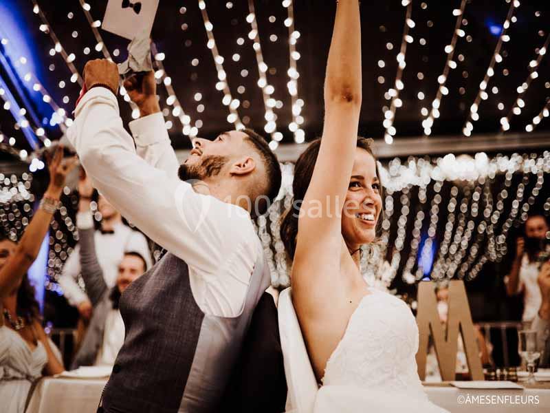 Un couple de mariés assis dos à dos lève des pancartes lors d'un jeu sous une décoration lumineuse.