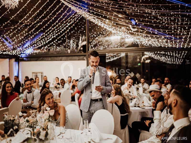 Un homme en costume lit un discours au micro devant des invités assis dans une salle décorée de guirlandes lumineuses.