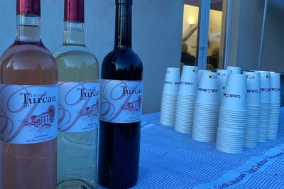 Salle avec un ancien pressoir en bois, des tonneaux, une table rouge avec bouteilles et des verres à vin.