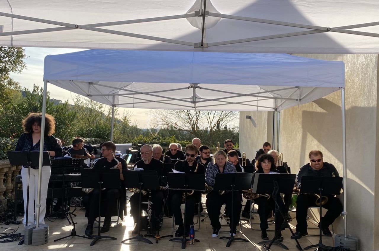 Un groupe de musiciens joue sous une tente blanche en extérieur, avec des pupitres et divers instruments.