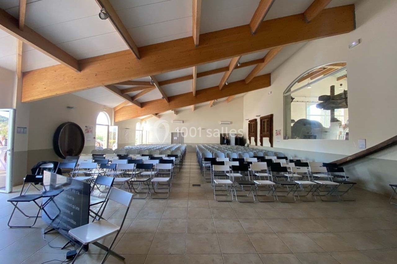 Salle lumineuse avec des rangées de chaises pliantes, plafond en poutres apparentes et grande baie vitrée.