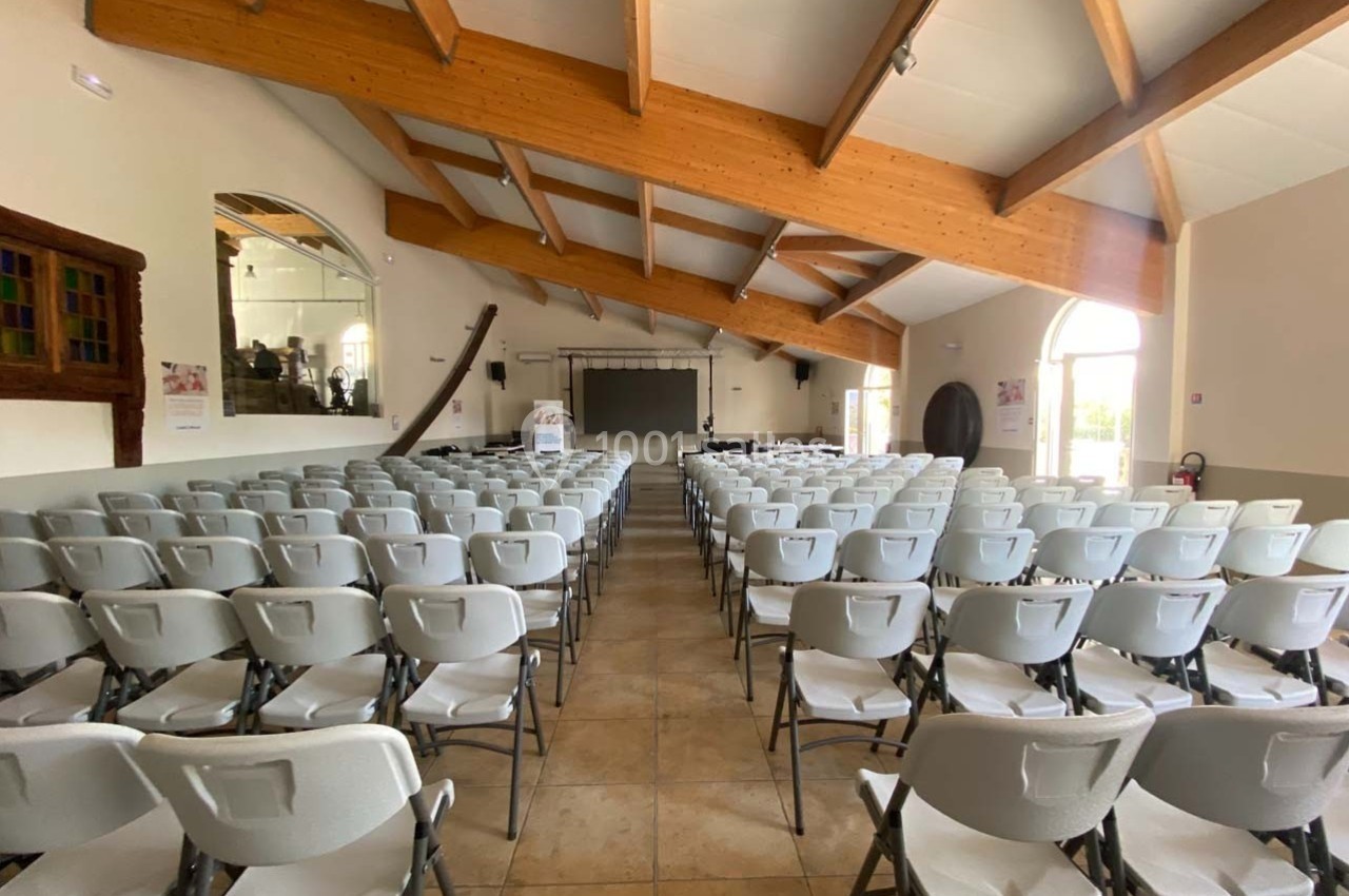 Salle lumineuse avec des rangées de chaises blanches alignées face à un écran et une scène, sous une charpente en bois.