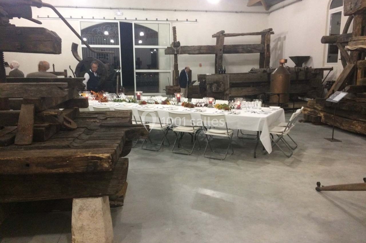 Salle avec des pressoirs anciens en bois, une longue table dressée pour un repas, et quelques personnes en arrière-plan.