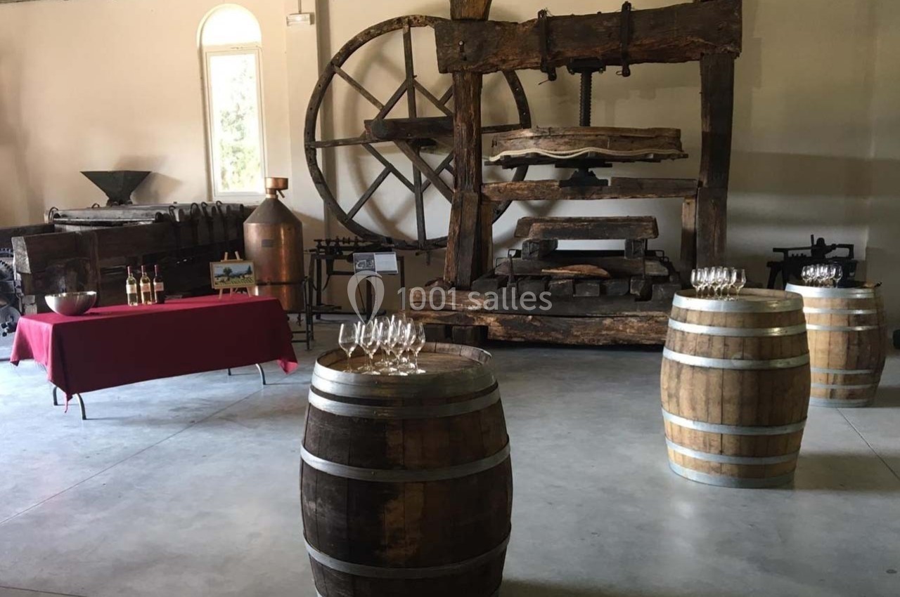 Salle avec un ancien pressoir en bois, des tonneaux, une table rouge avec bouteilles et des verres à vin.