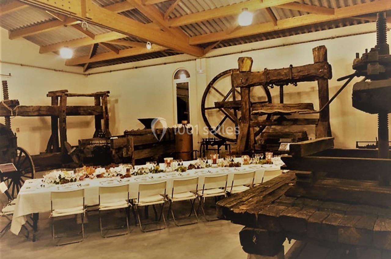 Salle avec des pressoirs anciens en bois exposés, une longue table dressée pour un repas au centre.