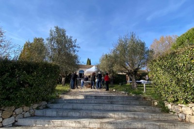 Salle avec un ancien pressoir en bois, des tonneaux, une table rouge avec bouteilles et des verres à vin.
