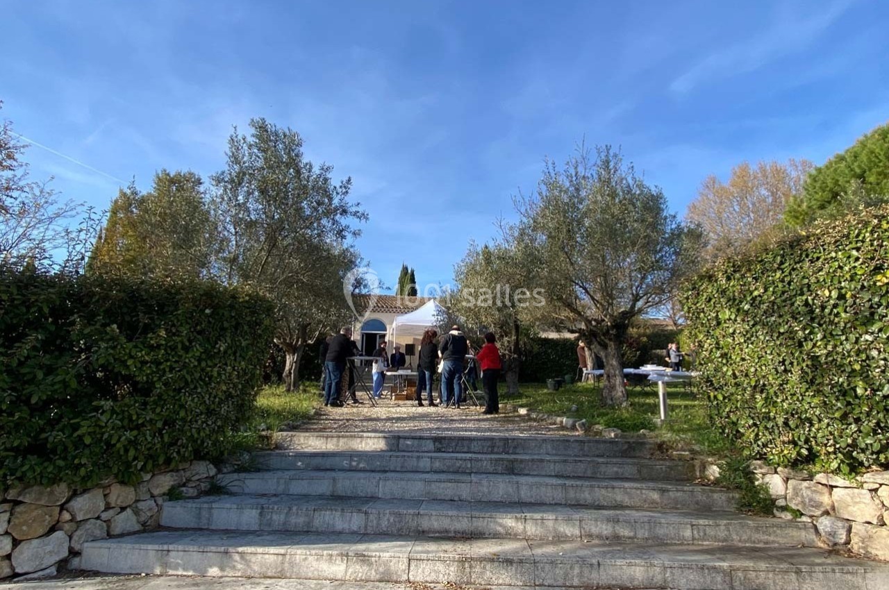 Groupe de personnes rassemblées dans un jardin devant une maison blanche entourée d'arbres et de haies.