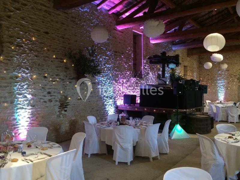 Salle en pierre avec tables dressées pour un événement, éclairage coloré et scène équipée pour la musique.