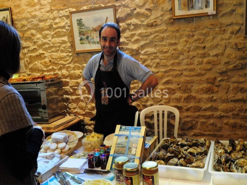 Un homme souriant derrière un stand de produits locaux, avec des huîtres, confitures et fromages exposés.