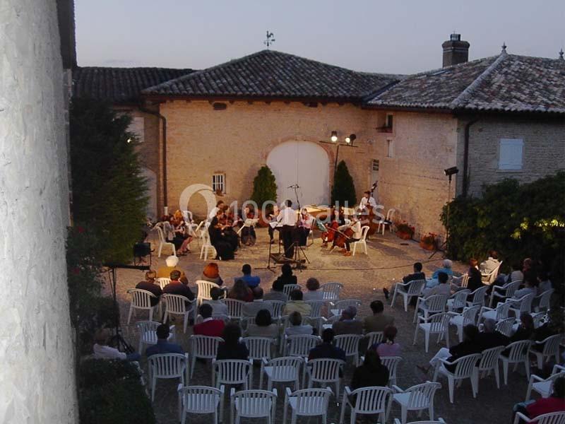 Concert en plein air dans une cour pavée au crépuscule, avec un orchestre et des spectateurs assis sur des chaises blanches.