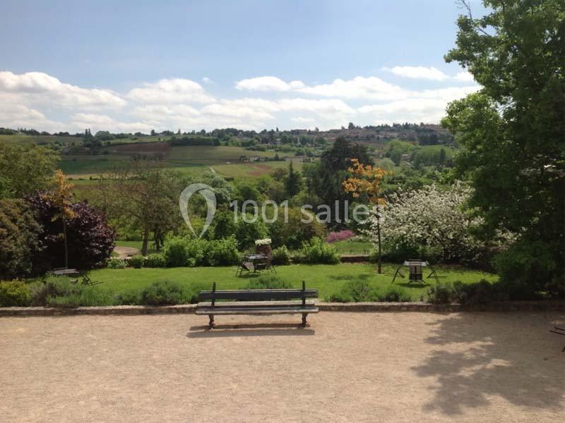 Vue d'un parc avec pelouse, arbres en fleurs, banc et collines verdoyantes sous un ciel partiellement nuageux.