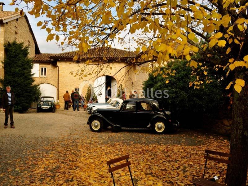 Voiture ancienne noire stationnée dans une cour pavée entourée de bâtiments en pierre et d'arbres aux feuilles jaunes.